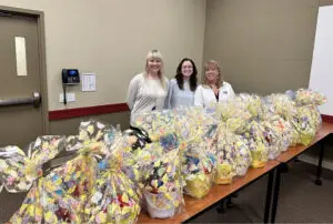 Three smiling women standing behind a table full of cellophane-wrapped Easter gift baskets.