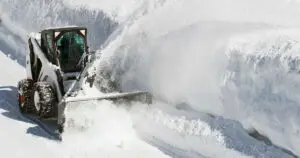 Skid steer loader with snow blower clearing deep snow from a road