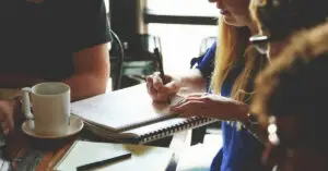 Woman writing in spiral notebook during team meeting at coffee shop table